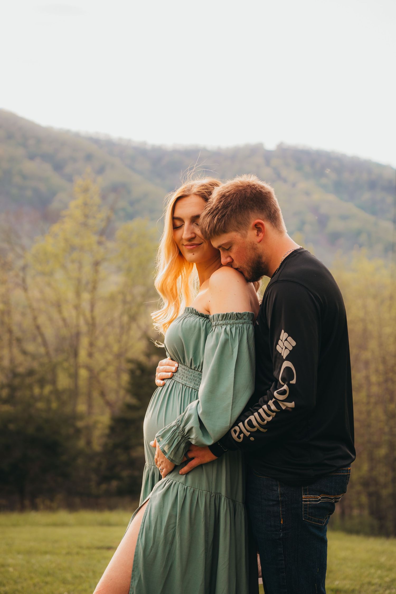 Loving maternity photo session in a green field with mountains in the background at sunset.