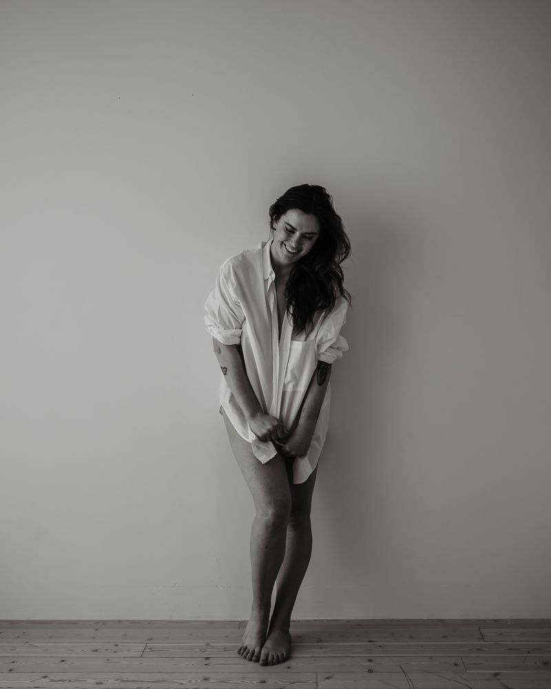 Black and white portrait of a woman in an oversized shirt posing against a minimalist wall.