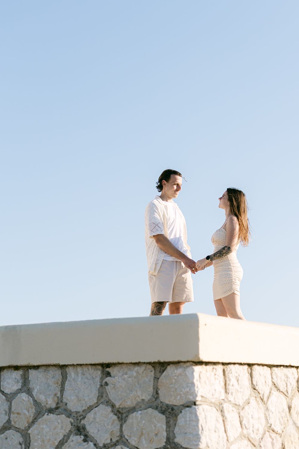 Couple in white summer outfits holding hands while standing on stone wall against clear blue sky.