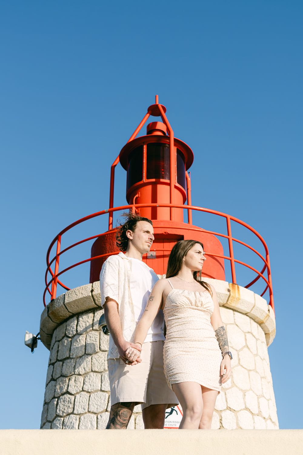 A couple poses together in front of a red-topped stone lighthouse tower under clear blue skies.
