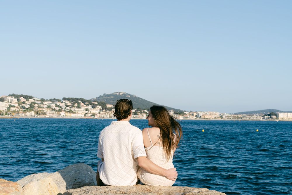 Couple sits together on rocky shore overlooking blue Mediterranean waters and coastal town.
