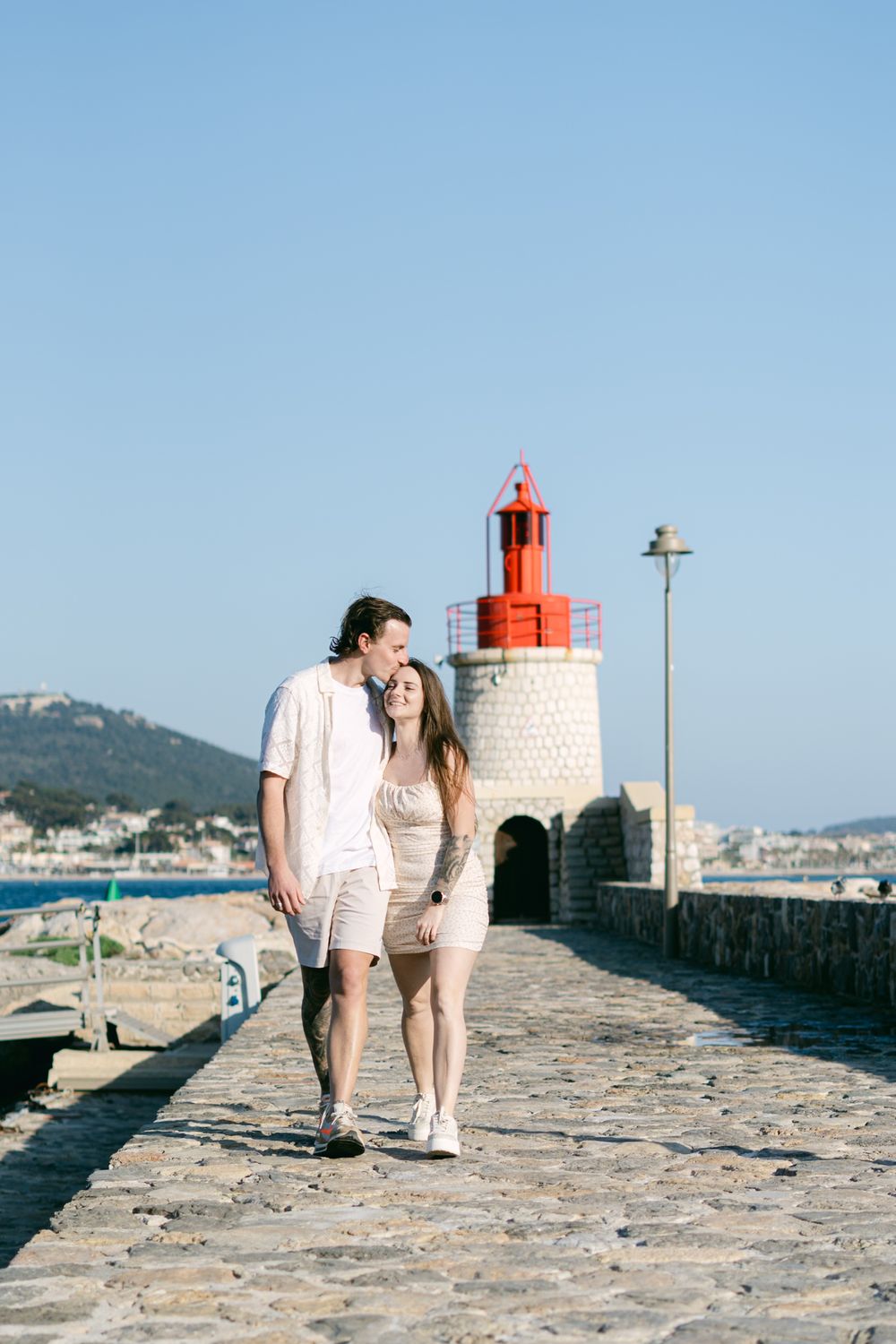 A couple walk closely together on a stone pier with a red and white lighthouse and coastal town in the background.