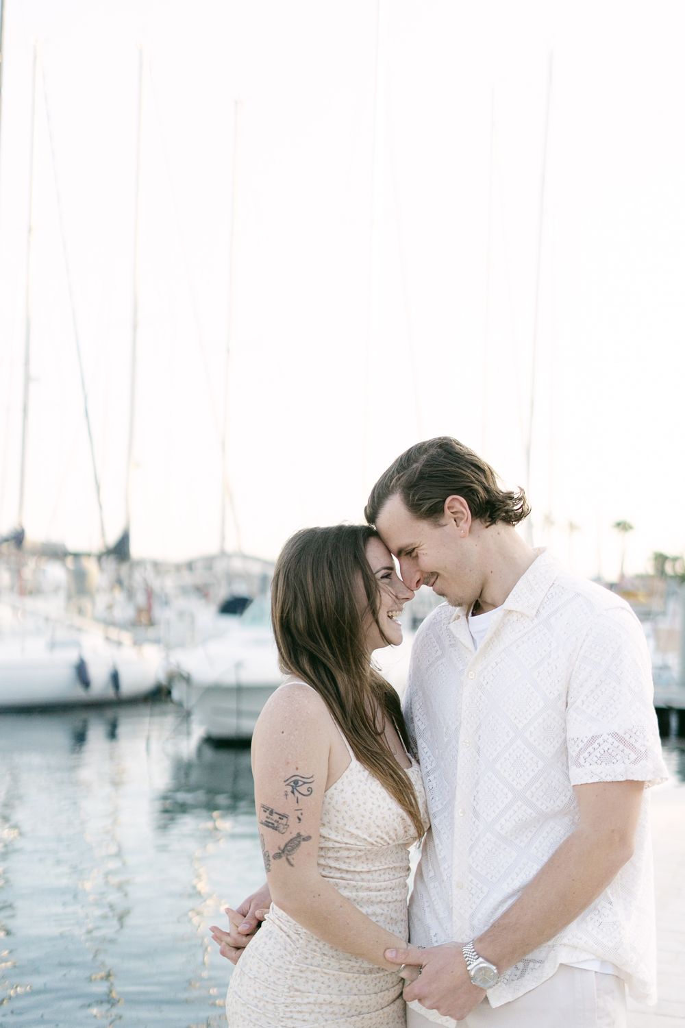 Romantic couple embraces near marina with sailboats in background during sunset.