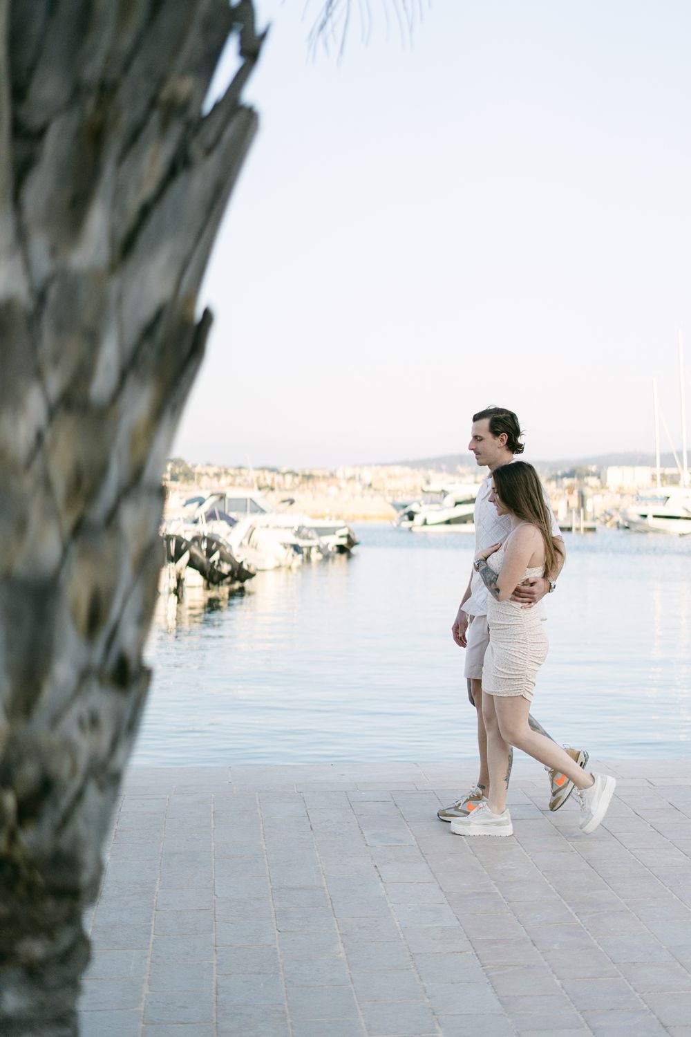 Couple walking hand in hand along waterfront promenade with marina and boats in background.