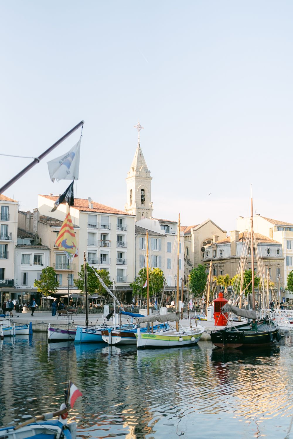 Boats docked in a Mediterranean marina with white buildings and a church bell tower in the background during sunset.