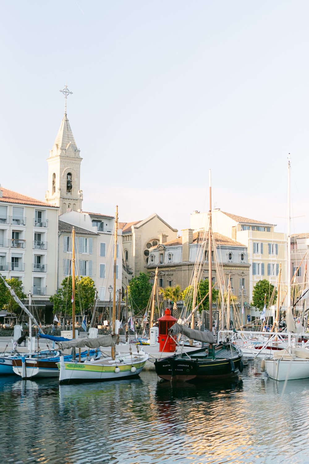 Sailboats docked in a marina with a white church steeple and Mediterranean style buildings along the waterfront.