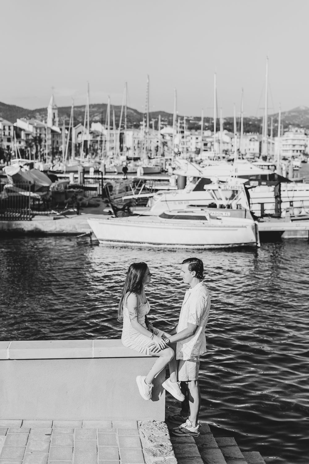 Two people sitting together on a dock overlooking a marina filled with sailboats and yachts at sunset.