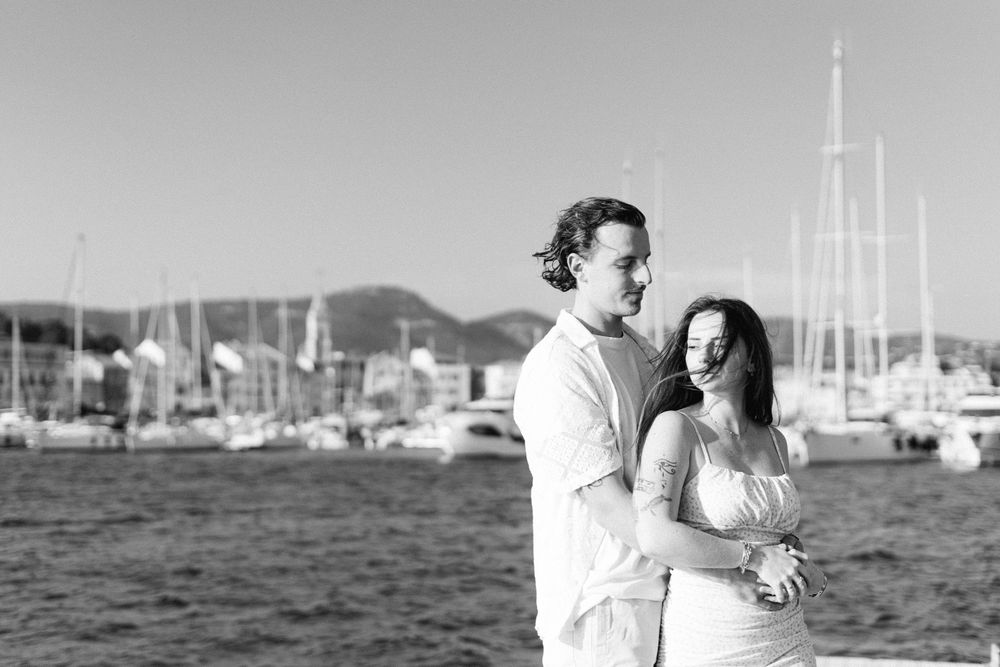 A black and white photo of a couple embracing by a marina with sailboats and mountains in the background.