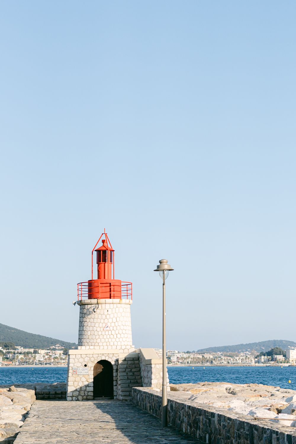 A red lighthouse tower with white stone base stands at the end of a stone pier overlooking a coastal town and blue sea.