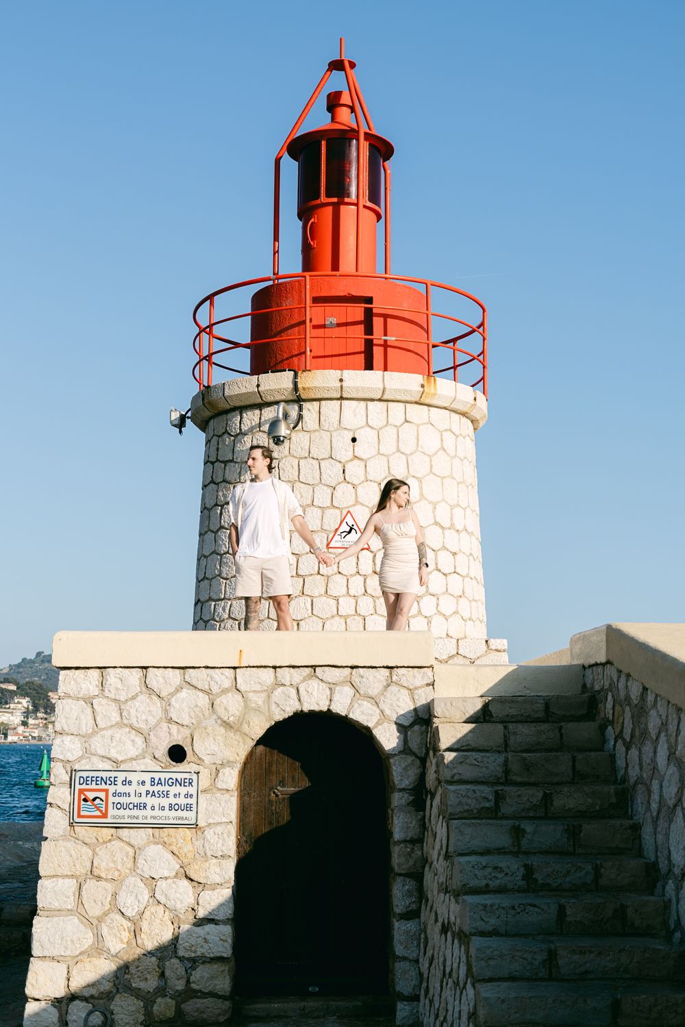 A lighthouse with a red top and stone base against a blue sky, showing a couple standing on its platform.