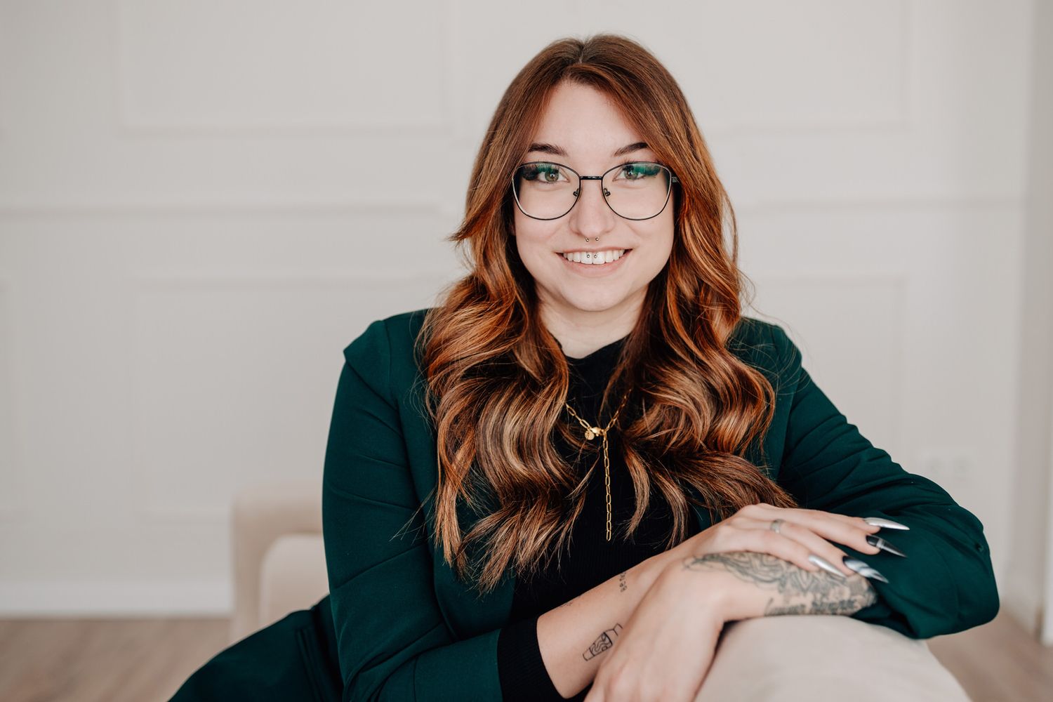 Someone in glasses and a dark green top smiles warmly while sitting against a light colored wall.Business Portrait Rheinmain Frankfurt