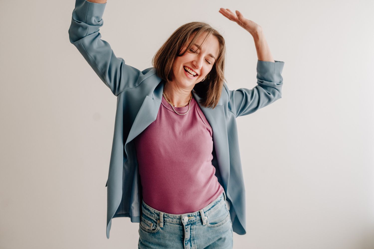 Someone in a blue blazer and pink top poses joyfully with raised arms against a white wall.