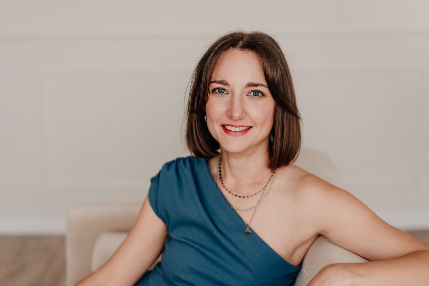 A smiling person in an elegant one-shoulder teal dress poses for a portrait against a neutral background.
