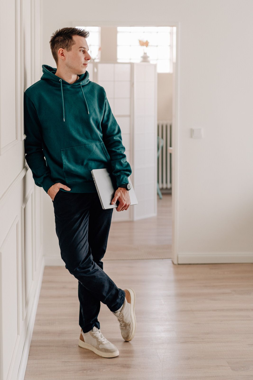Someone in a dark green hoodie and black pants leans against a white wall while holding a laptop in a minimalist interior.Business Portrait