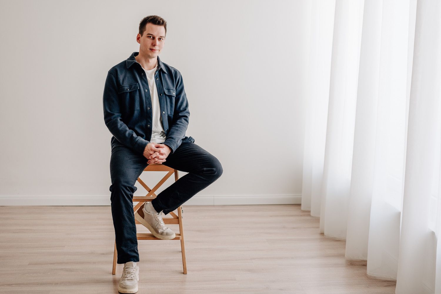 Professional in navy jacket and jeans sitting casually on wooden chair against white curtained backdrop.