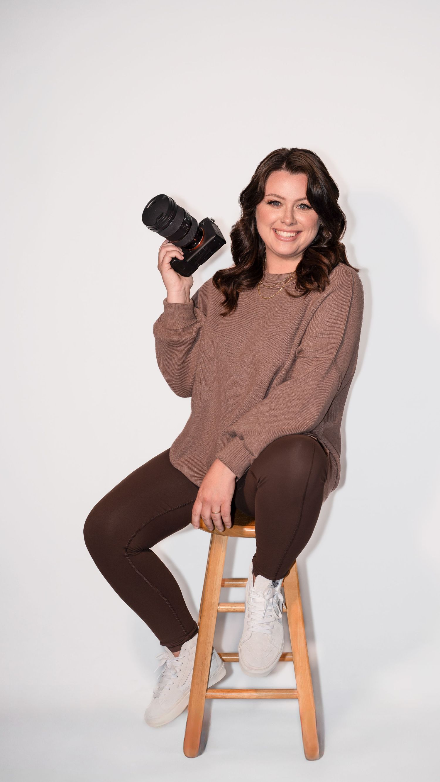Professional photographer Maggie Martinez in brown sweater and pants sits casually on wooden stool holding camera against white background.