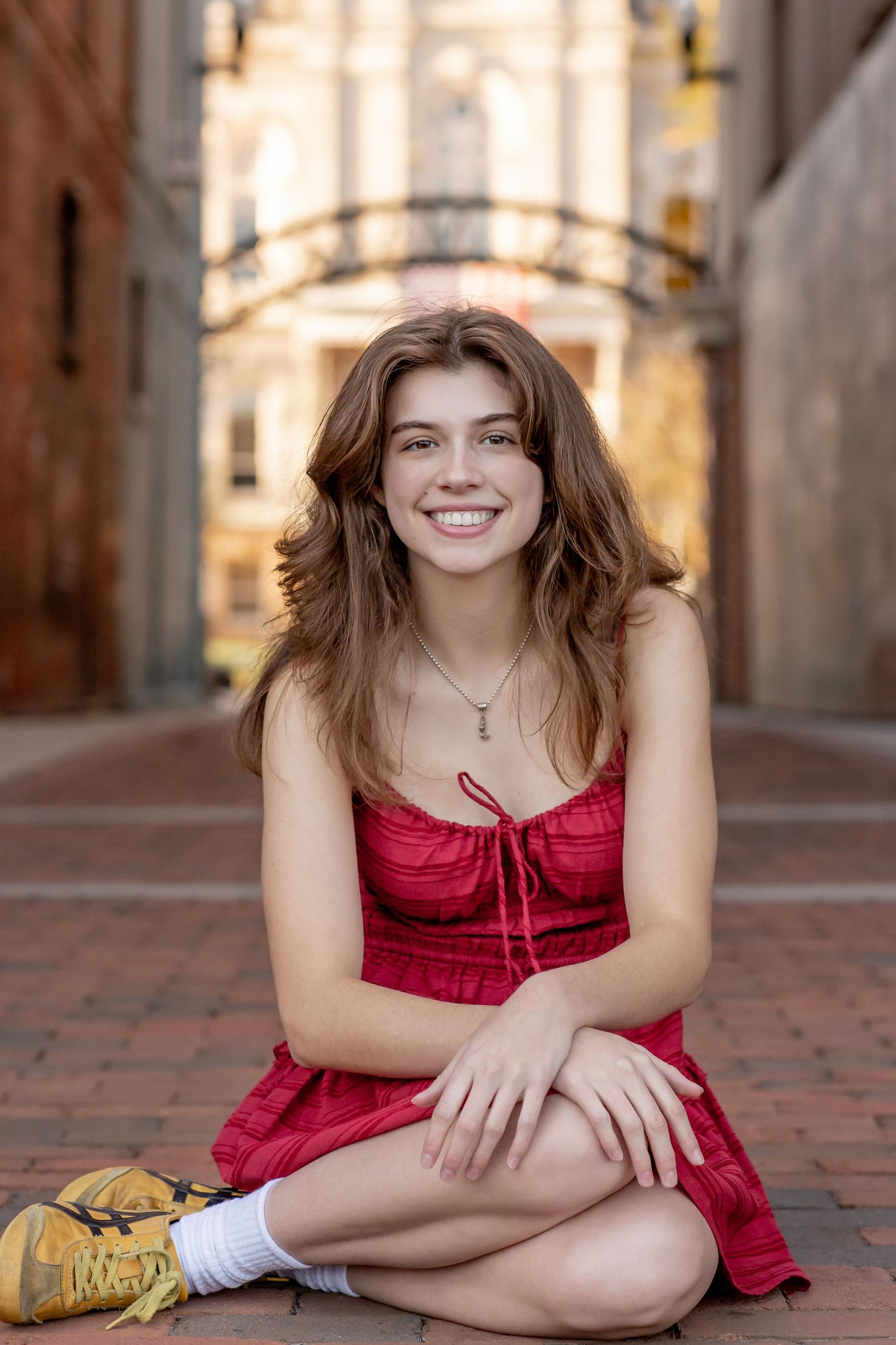Young woman in red dress sitting cross-legged on brick pathway smiling outdoors.
