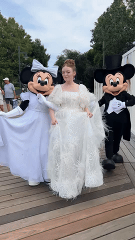 Mickey Mouse characters in wedding attire pose with figure in white feathered gown on wooden deck outdoors.