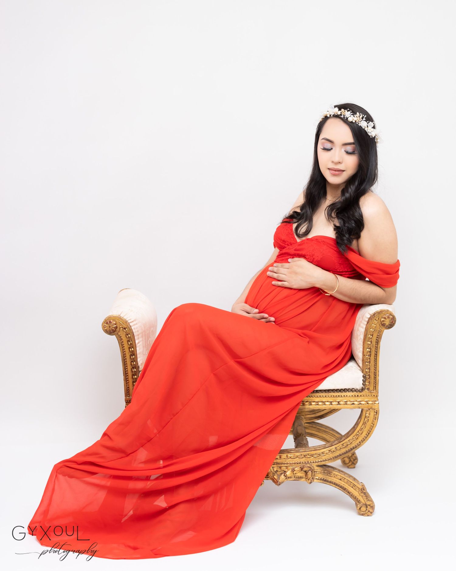 Expectant mother in red off-shoulder gown poses on vintage gold chair.