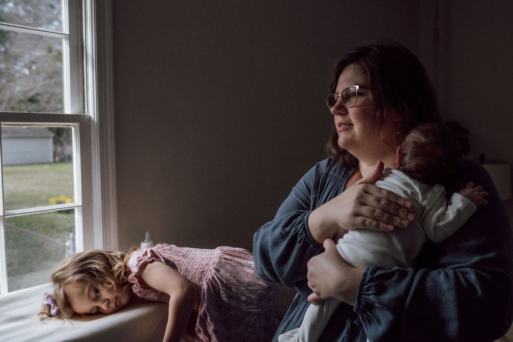 A quiet moment by the window shows a child sleeping on the windowsill while someone tenderly holds an infant nearby. in home newborn photo