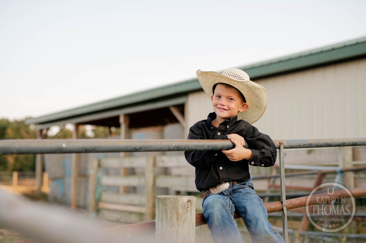 Young cowboy in black shirt sits on metal fence rail against barn backdrop.