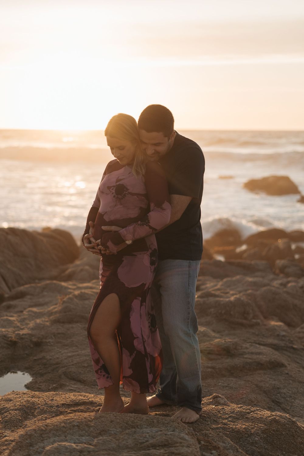 Silhouettes of a couple and child embracing on rocky beach at sunset with ocean waves in background.