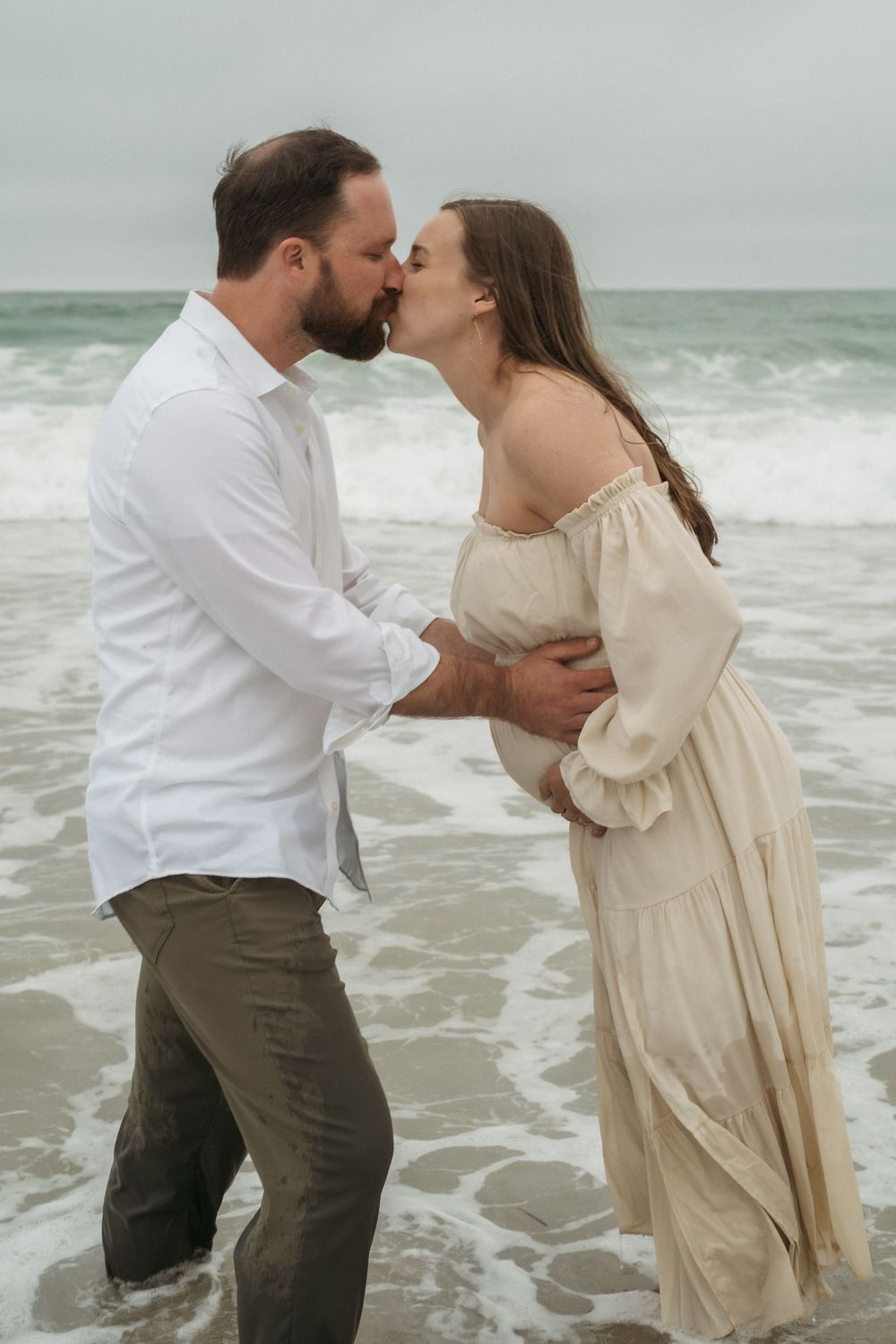 Romantic couple in light beige and white clothing share intimate moments while standing in ocean waves at the beach.