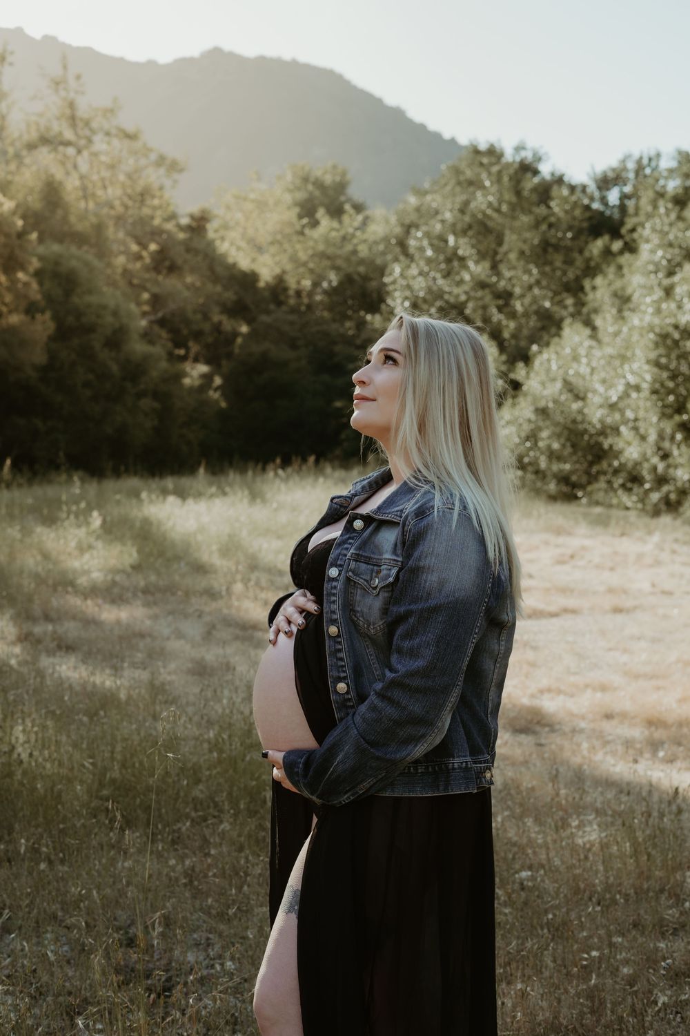Outdoor maternity portrait with woman in denim jacket gazing upward against mountain and sunset landscape.