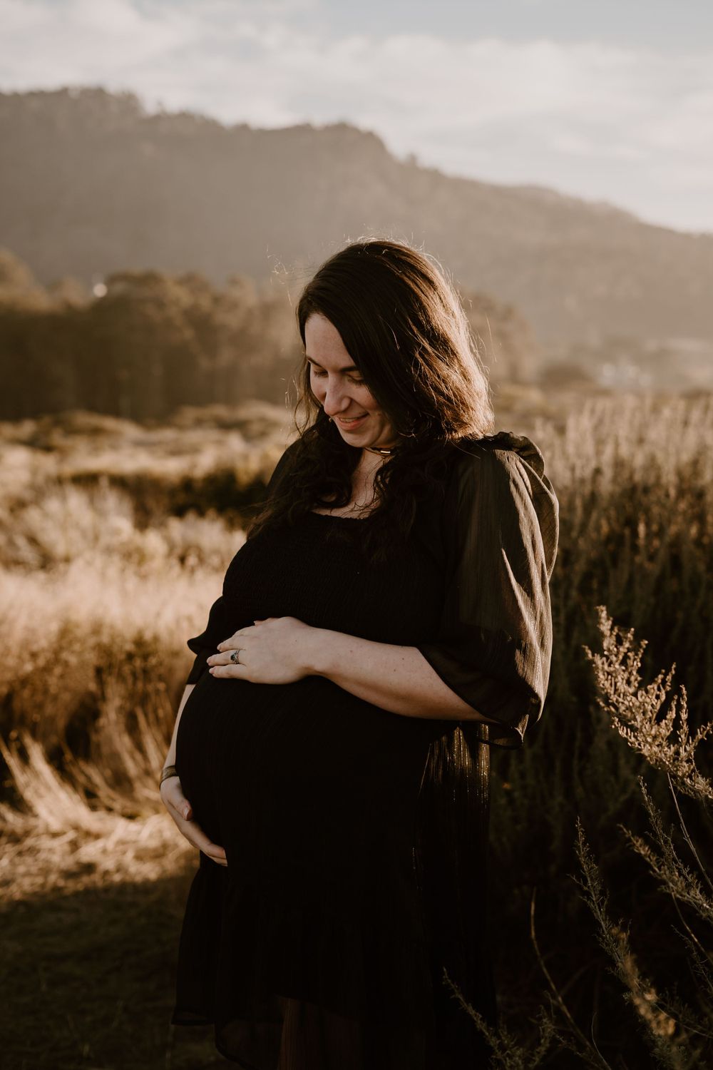 Expectant mother in black dress smiles while touching her belly during golden hour with hills in background.