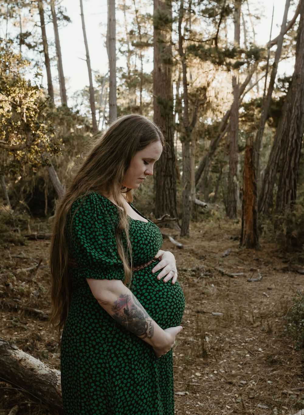 A pregnant woman in a green polka dot dress stands in a forest during golden hour with sunlight filtering through trees.