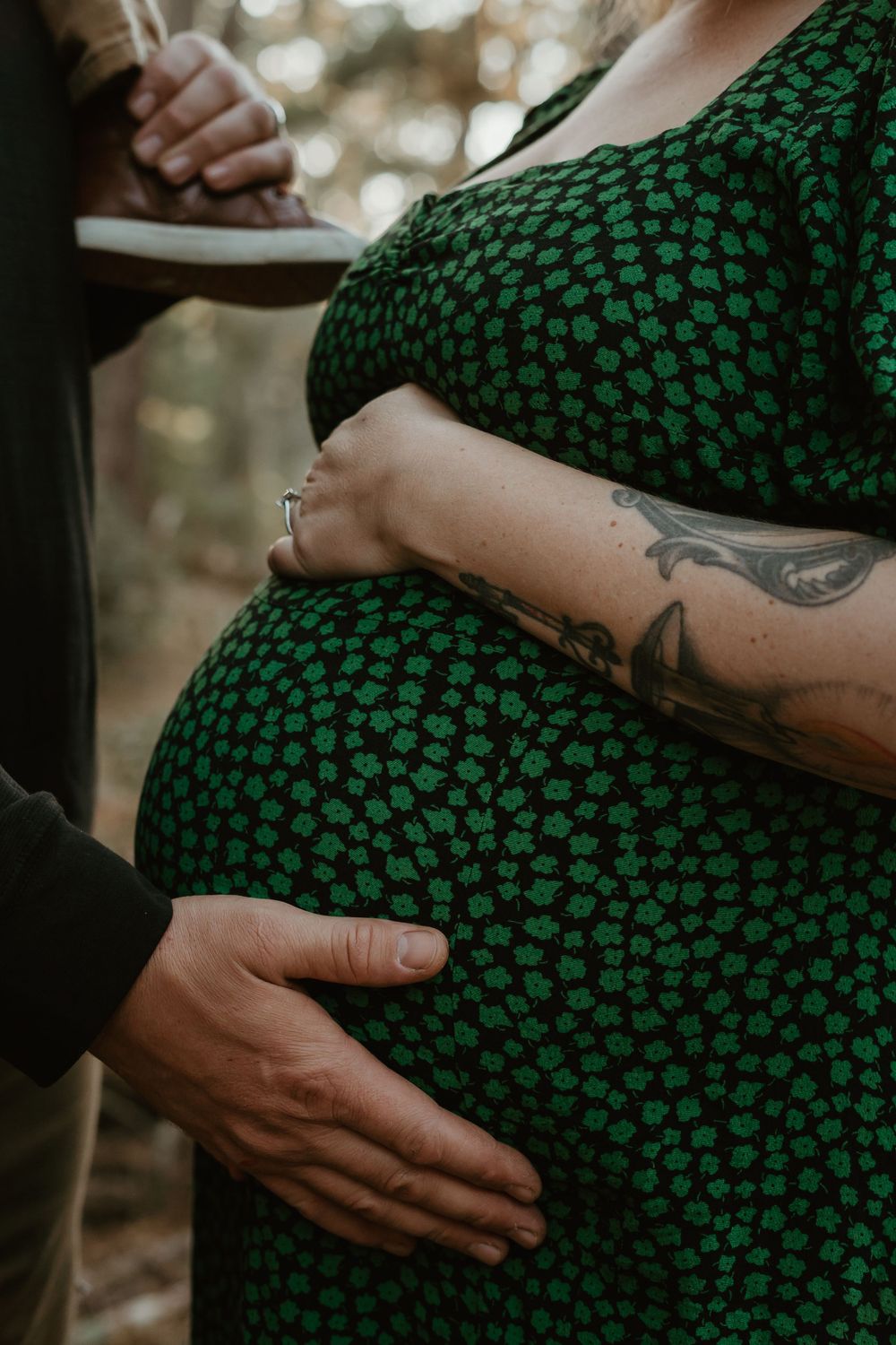 Close-up of a pregnant belly in a green patterned dress with hands tenderly touching and a visible tattoo on the arm.