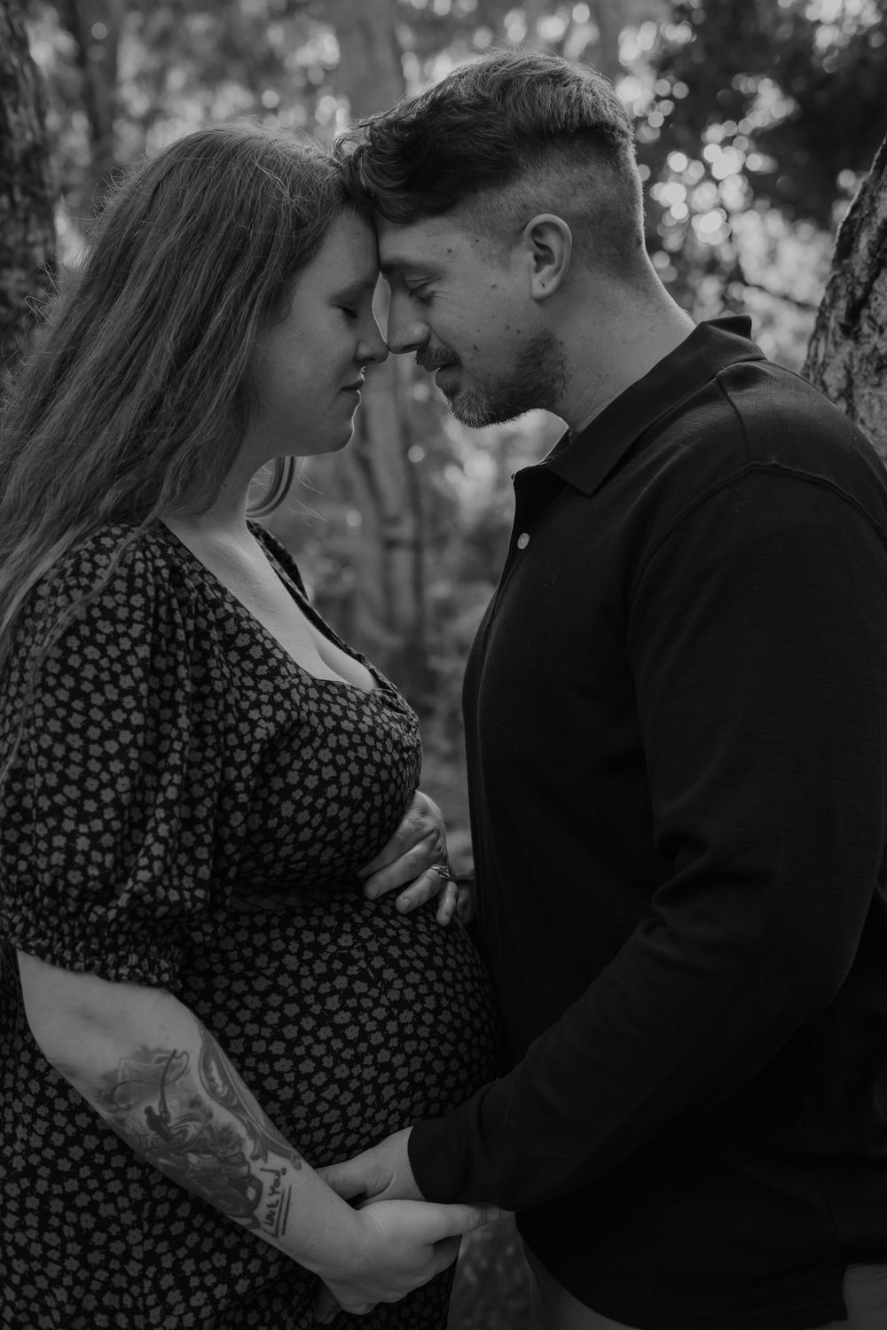 A romantic black and white maternity photo shows an intimate couple embracing in a forest setting.