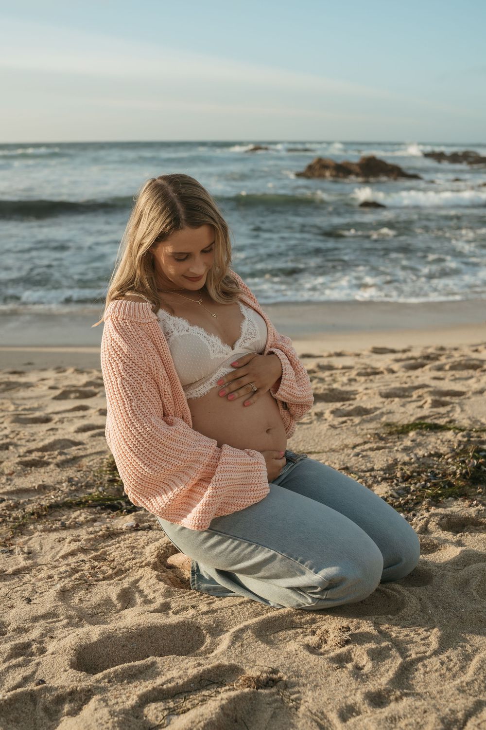 A mother breastfeeding her baby on a sandy beach with ocean waves in the background during golden hour.