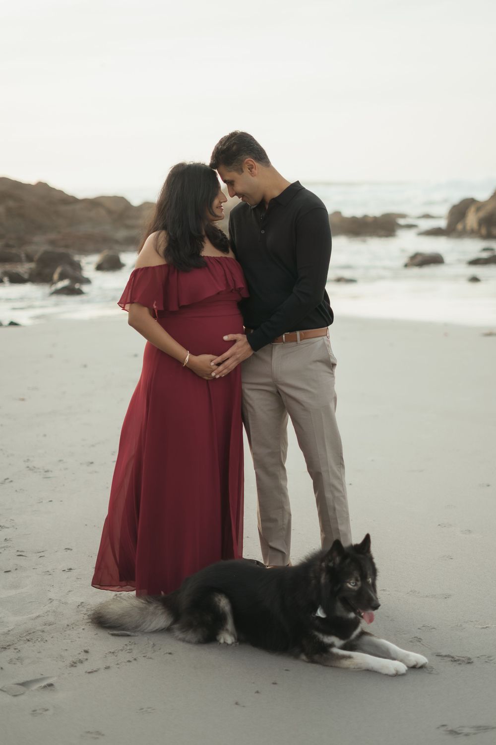 A couple embraces on a misty beach with their black and white dog sitting beside them on the sand.