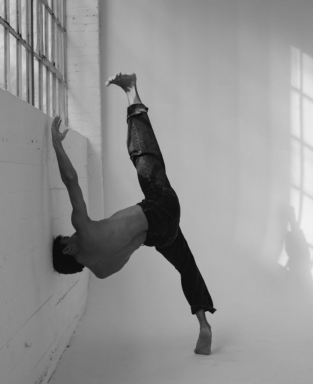 Athletic dancer performs dramatic wall-supported pose in black and white.