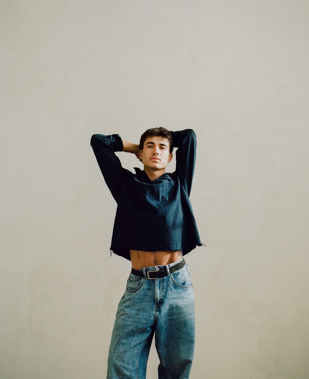 Model wearing a cropped navy hoodie with blue jeans in a minimal studio setting against a beige background.