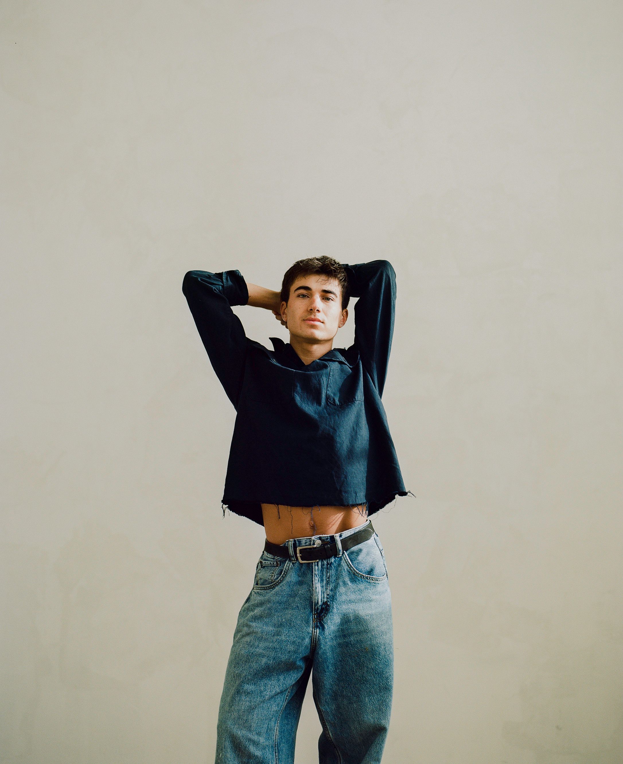 Model wearing a cropped navy hoodie with blue jeans in a minimal studio setting against a beige background.