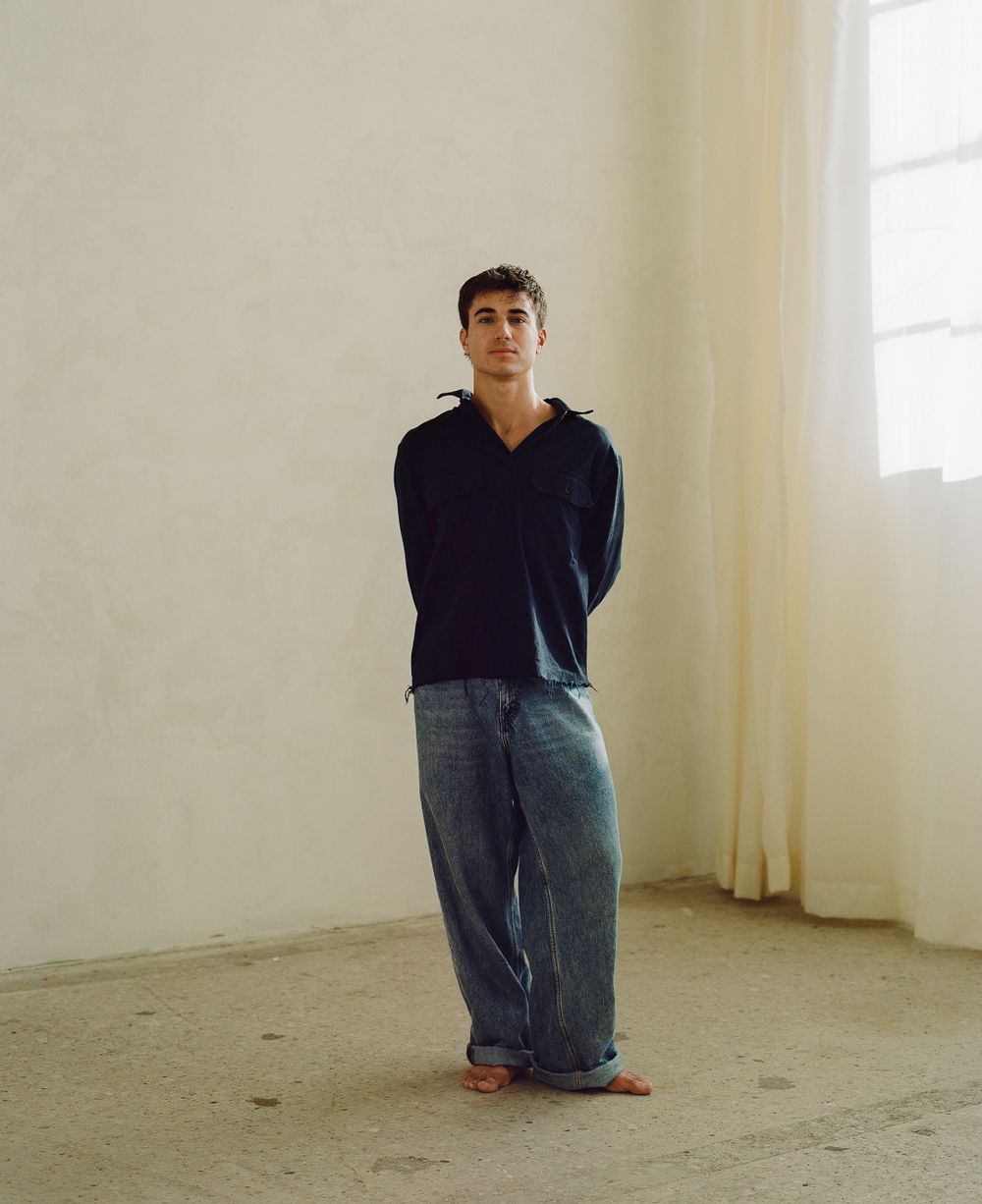 Person in navy hoodie and loose-fit jeans standing against white wall in minimalist interior.