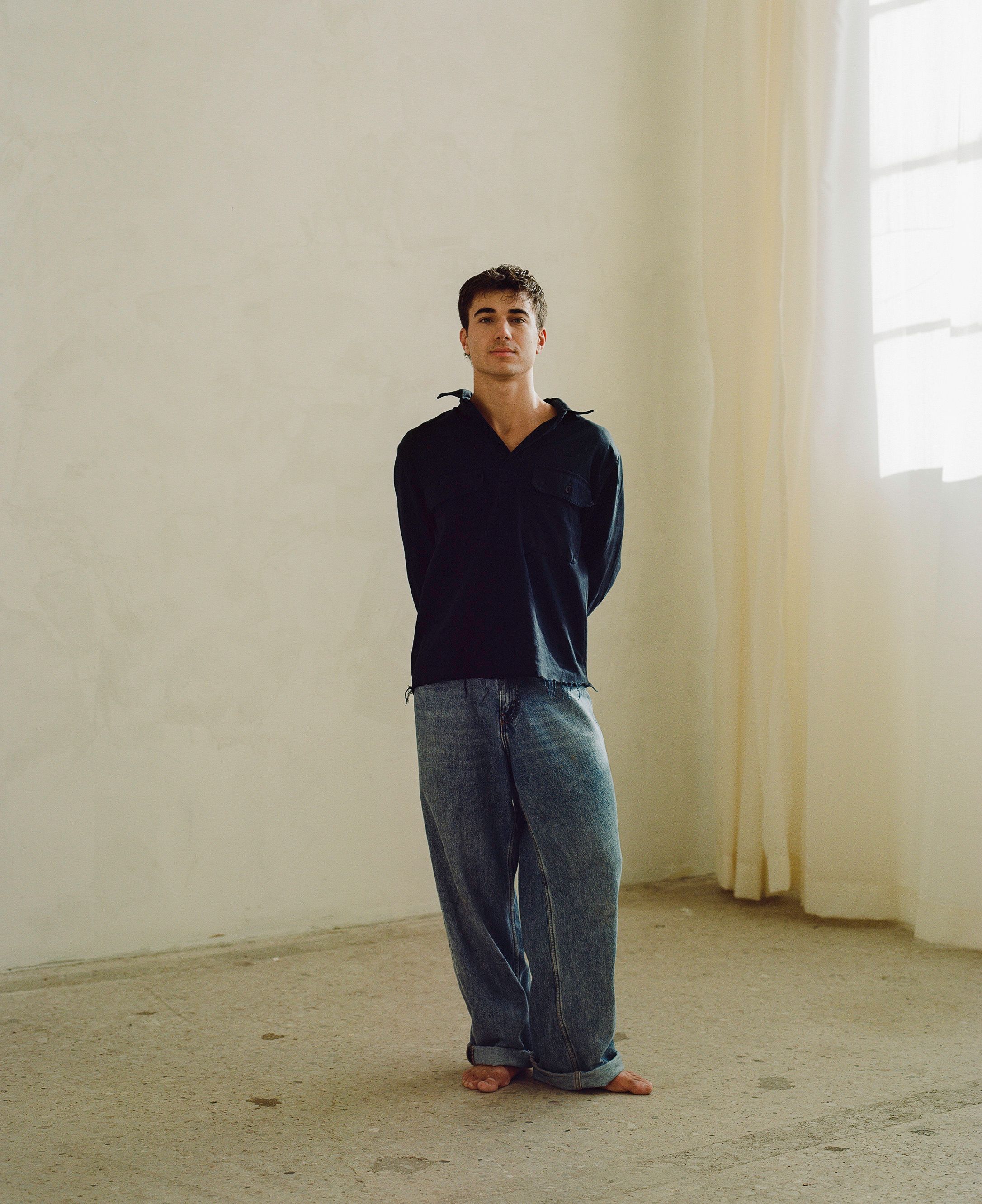Person in navy hoodie and loose-fit jeans standing against white wall in minimalist interior.