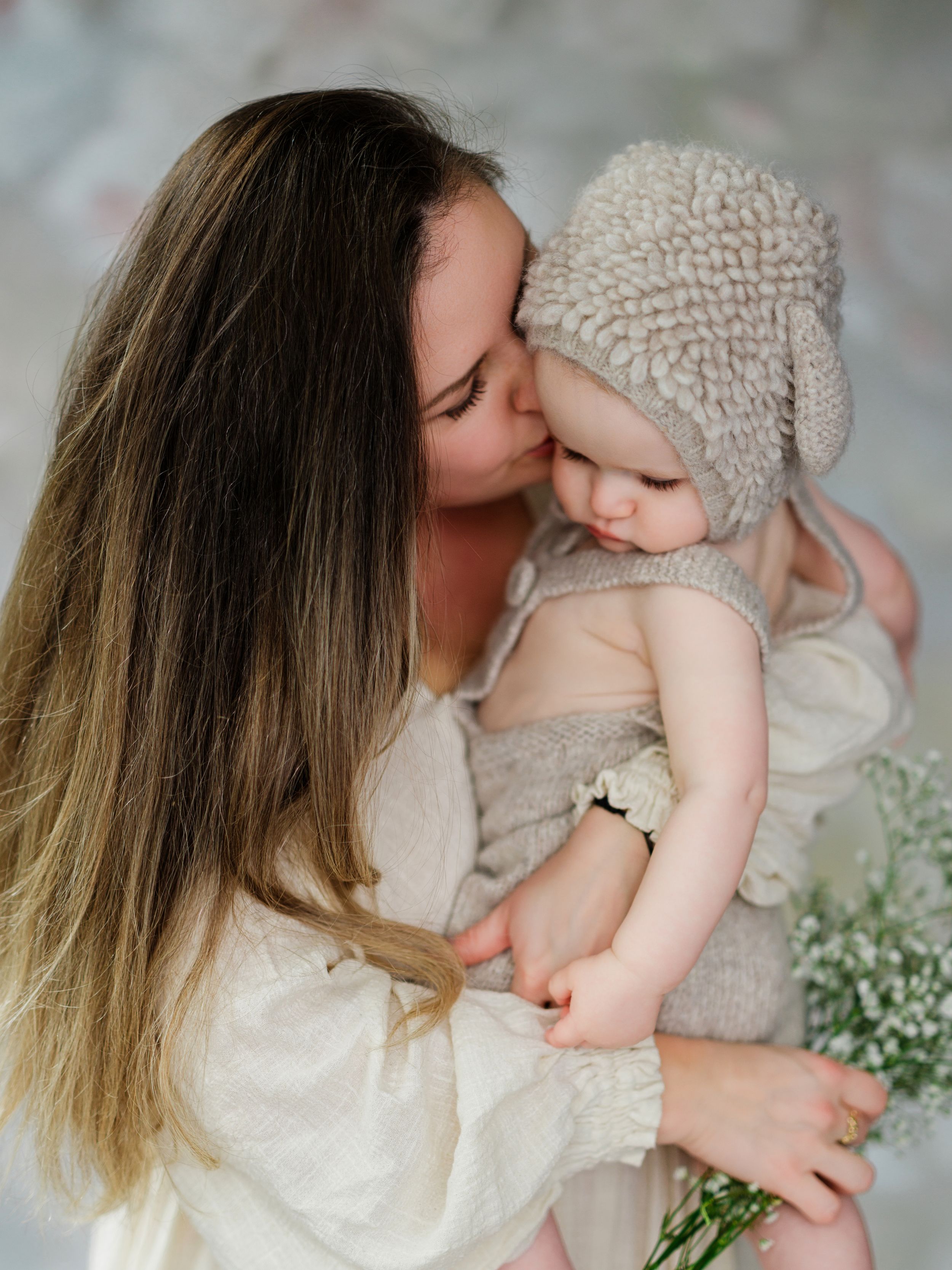 A tender moment captured as a mother in white dress kisses her baby wearing a knit bonnet among delicate flowers in Bellevue studio.
