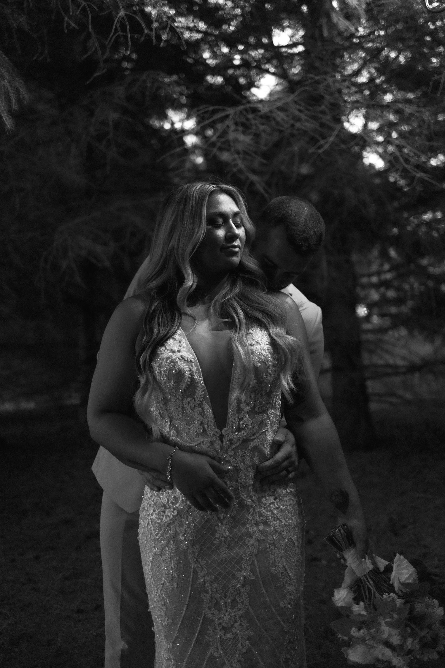 A romantic black and white wedding photo showing a couple embracing in a forest setting with the bride wearing a lace gown.