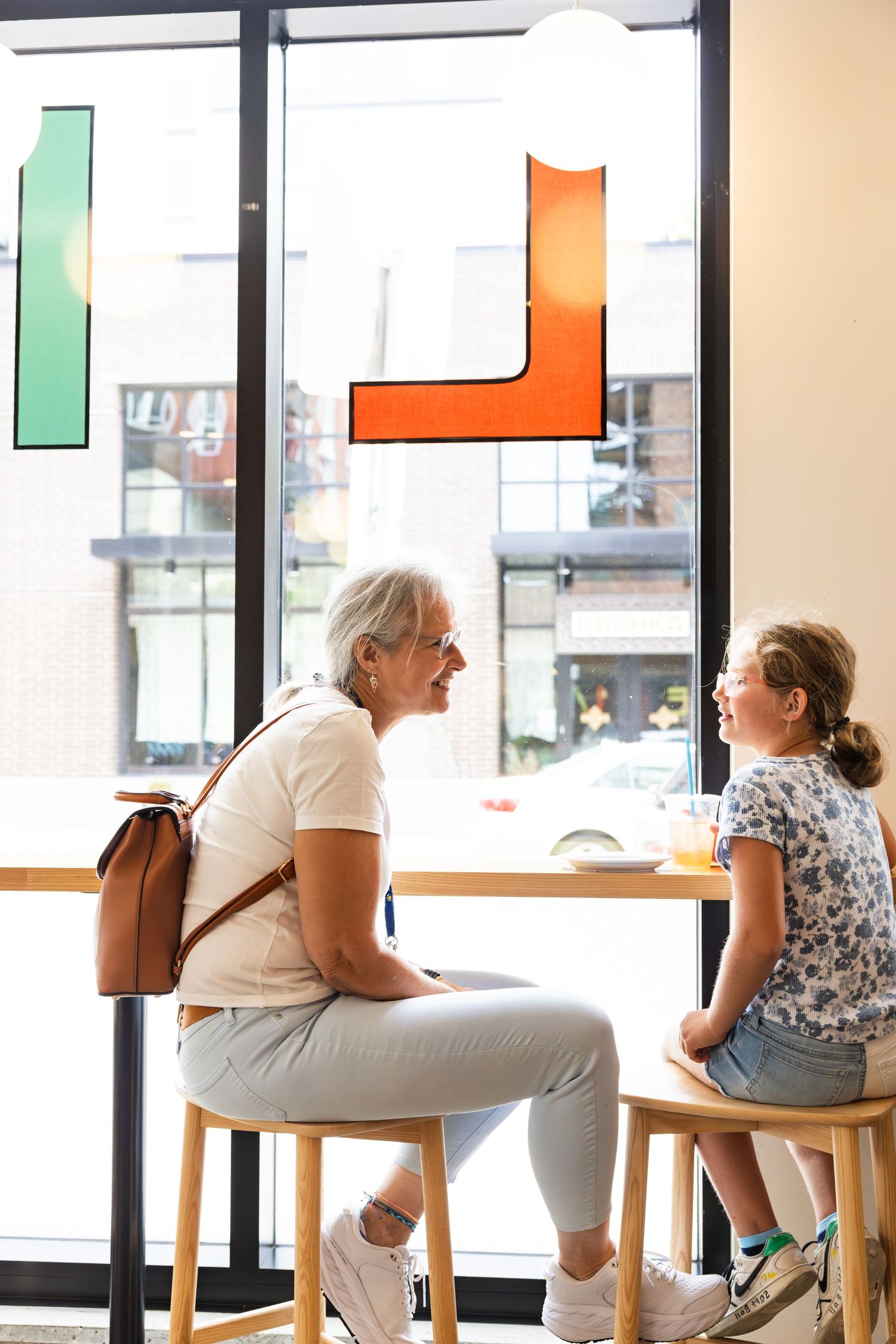 Two people sit and talk at a wooden table by a window in a bright modern cafe setting.