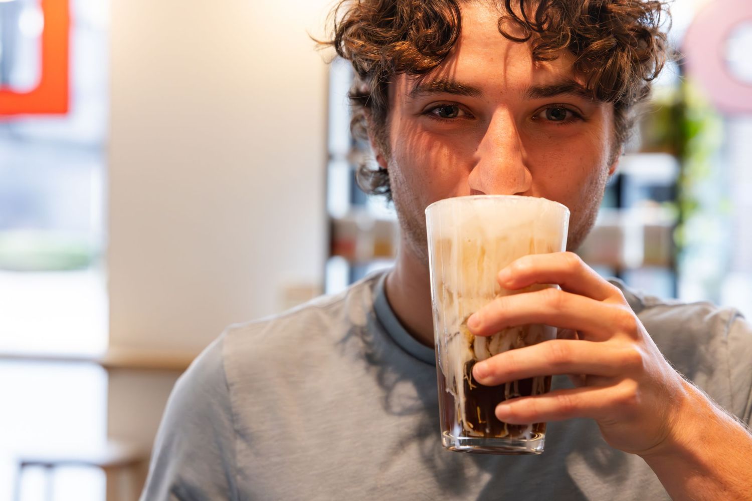 Someone with curly hair drinks a creamy cold brew coffee with foam at a cafe.