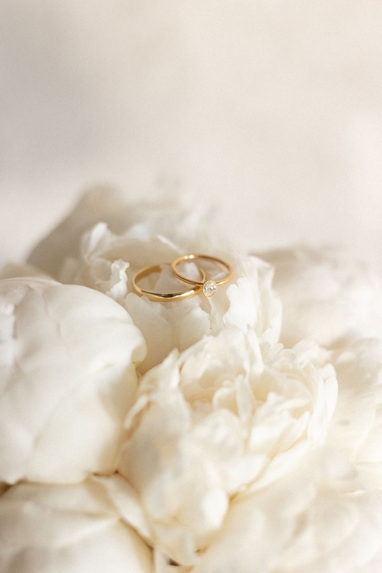 Two golden wedding rings nestled among delicate white peony petals in soft lighting.