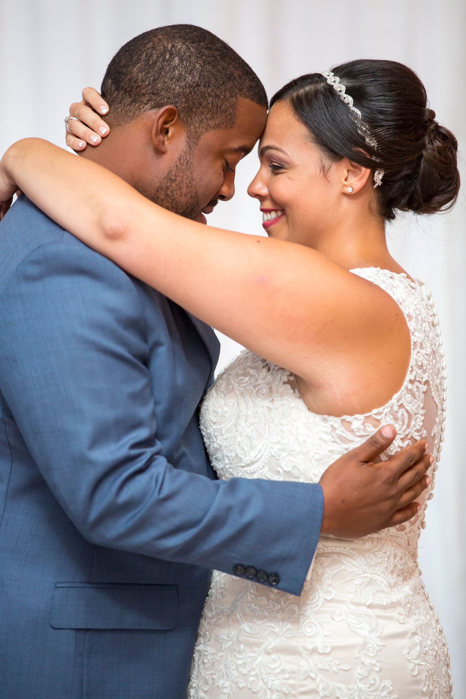 A couple shares an intimate moment in wedding attire, with the bride in a lace gown and groom in a blue suit.