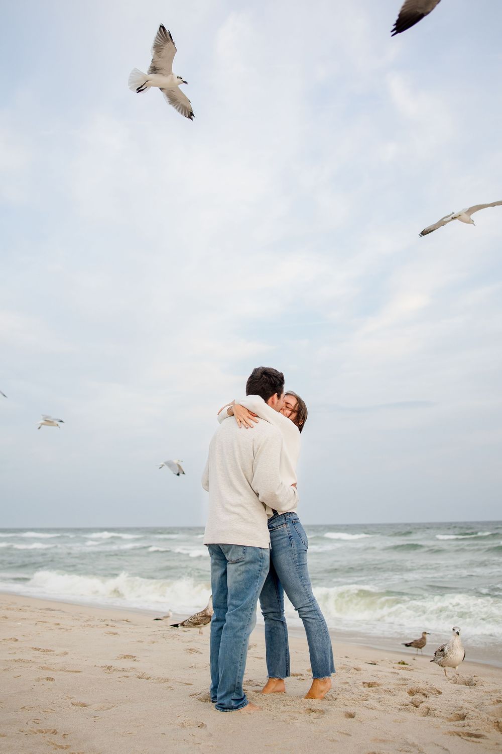 A couple embraces on a beach while seagulls soar overhead against a cloudy sky and ocean waves.