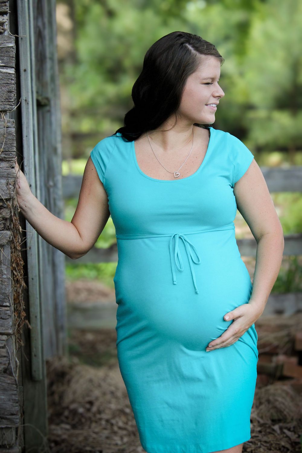 An expectant mother in a turquoise maternity dress poses outdoors near a rustic wooden structure.