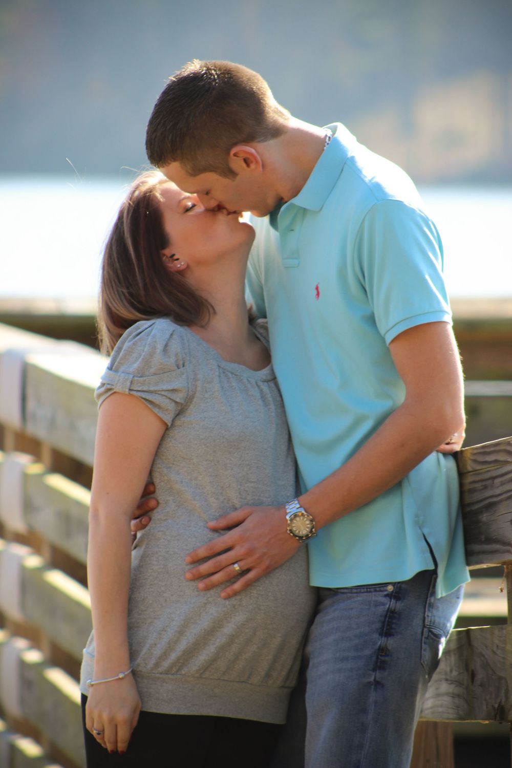 A romantic couple in casual attire share tender kisses while standing by a wooden railing outdoors.