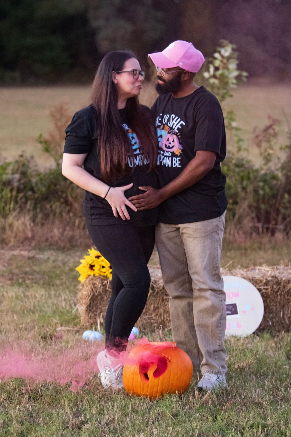 A couple poses together in matching t-shirts next to a pink smoke bomb and orange pumpkin in a grassy field.
