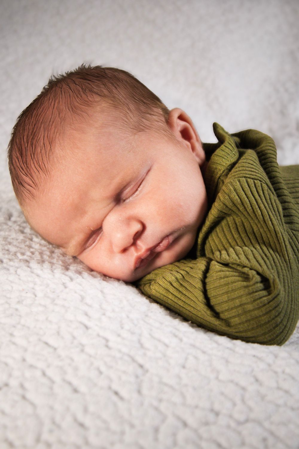 A sleeping newborn baby wrapped in an olive green swaddle blanket rests peacefully on white textured fabric.